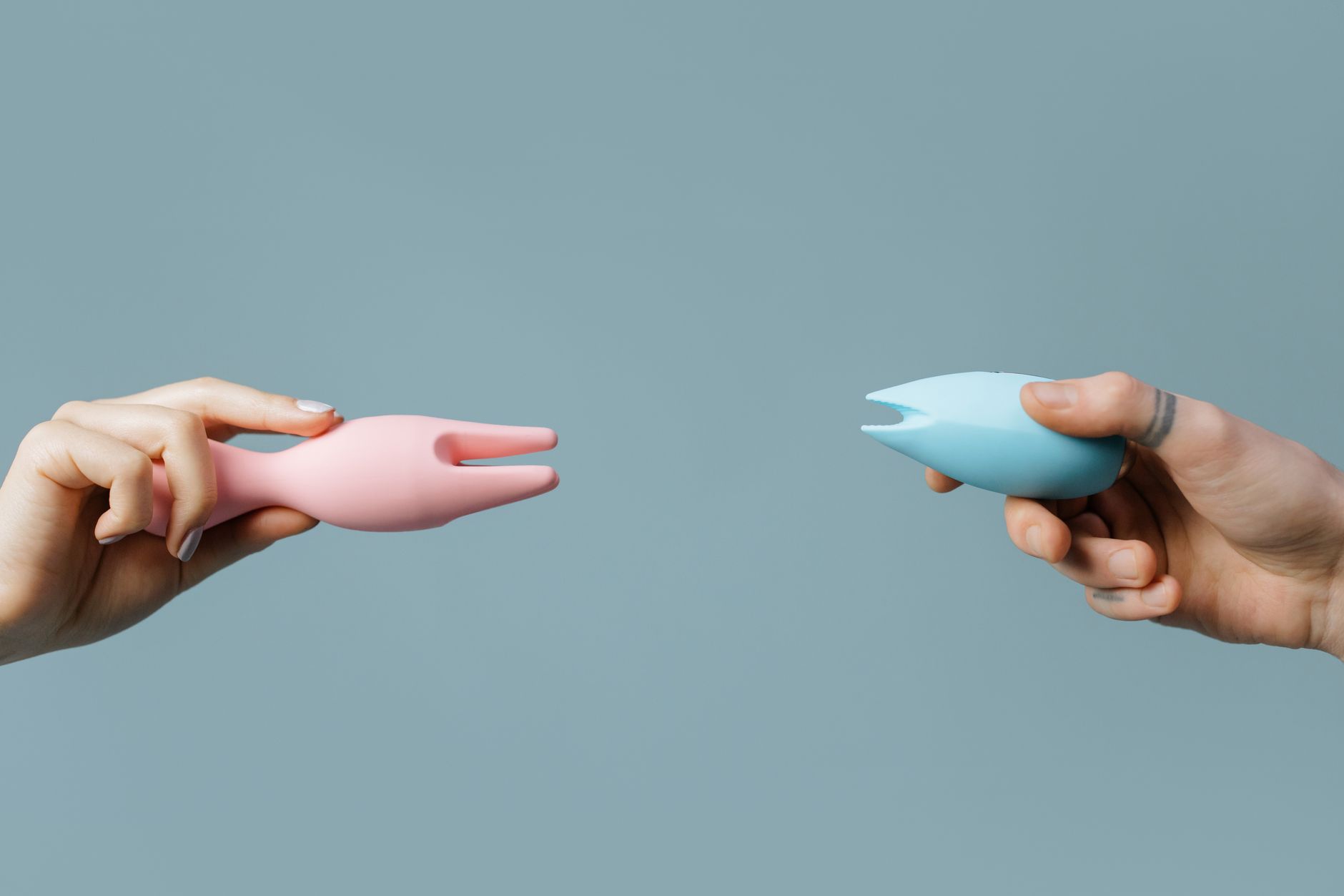 Duas mãos segurando vibrador rosa e azul contra um fundo pastel