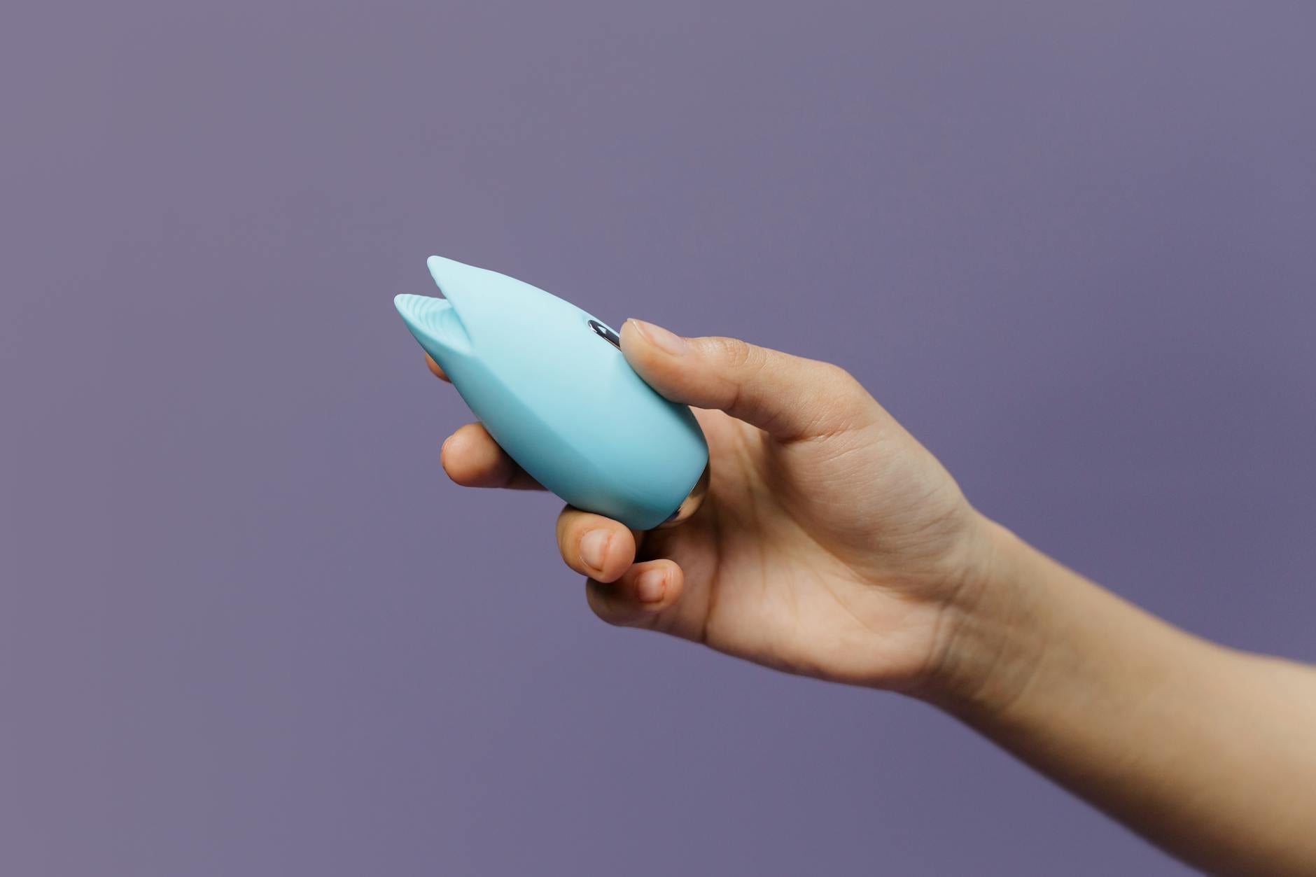 Um vibrador de silicone azul sendo segurado na mão contra um fundo roxo sólido, promovendo o auto-amor e a sexualidade